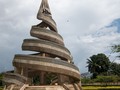 The Reunification Monument, Mamfé Cameroon. Commissioned by President Ahmadou Ahidjo in the 1970s the design and its creators nod to the transition from colonial rule to independence. The three people who designed it each played a role in the creating of modern Cameroon. They were Cameroonian sculptor and artist Gédéon Mpondo who had studied at l'Université des Beaux-arts in Paris in the late 1960s, the French architect Armand Salomon who played a significant role in creating the modern urban landscape of independent Cameroon and was the ‘first foreigner to­ be a licensed archi­tect in Cameroon’* and Cameroonian architect Engelbert Mveng. The striking sculpture, a spiral of multi-shaded concrete, is said to represent reunification, the merging of what is described as the ‘two Cameroons’, French and British. Some have written that the spirals are actually two snakes whose heads merge together. Out of shot is the image of an gnarled elderly man surrounded by children - holding a flame, presumably lighting the way to the future. Image Steve Mvondo CC BY SA 3.0 __________ #ArchitectureNow All materials presented on this site are Ⓒcopyrighted and owned by the creators listed above.